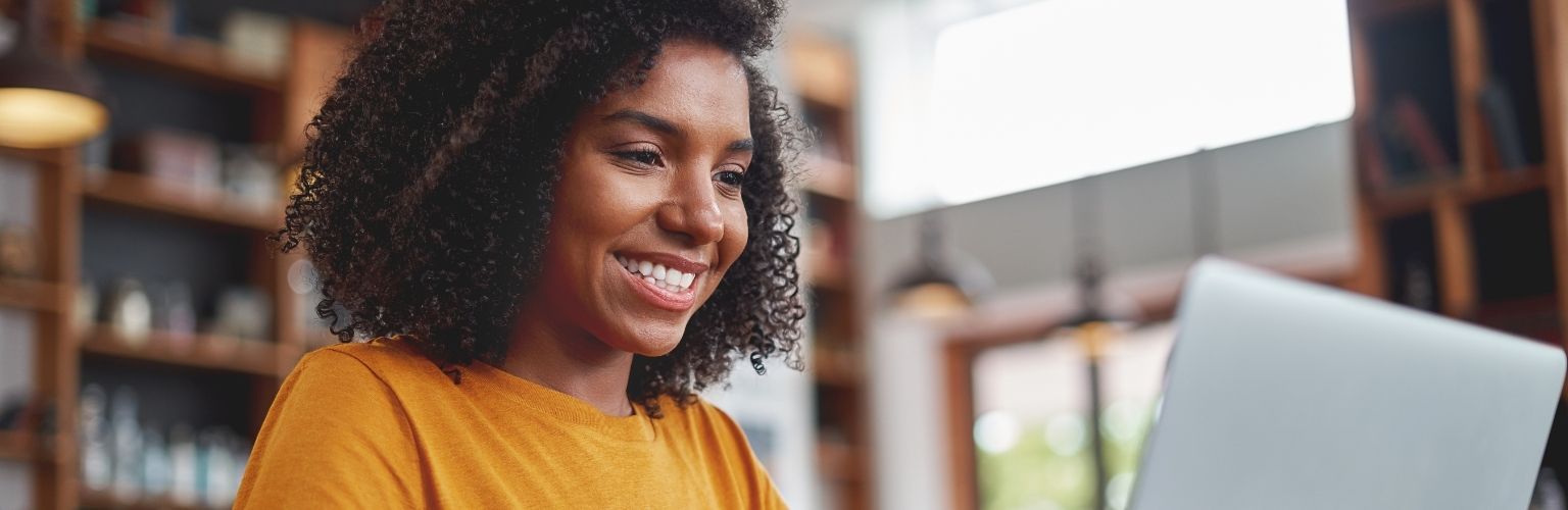 A woman with curly hair and a yellow top smiles while looking at a laptop in a cozy interior space.