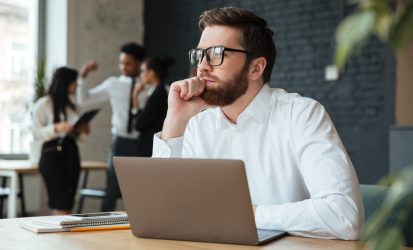 Image of concentrated young caucasian businessman sitting indoors using laptop computer. Looking aside.