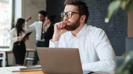 Image of concentrated young caucasian businessman sitting indoors using laptop computer. Looking aside.